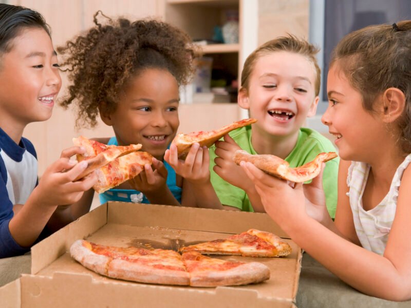Four young children indoors eating pizza smiling