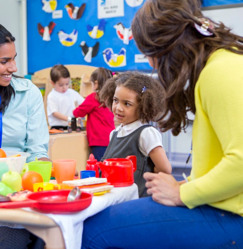 Teachers playing with plastic kitchen toys with their nursery students in the classroom.